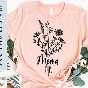 May include: A light pink t-shirt featuring a black floral bouquet design with the word "Mema" in a script font. The shirt is made of soft material and is perfect for everyday wear.
