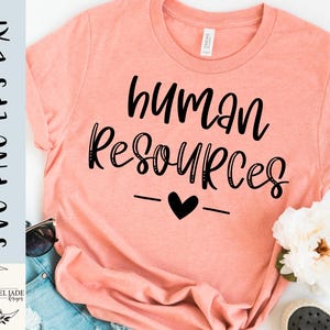 May include: Peach-colored t-shirt with the words "human resources" in black script, a black heart, and two horizontal lines. The shirt is displayed with sunglasses, denim shorts, and a white flower.