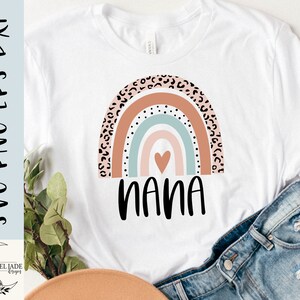 May include: A white t-shirt with a rainbow design featuring a heart in the center and the word "nana" underneath. The rainbow is made up of pink, orange, blue, and green stripes with a leopard print pattern on the outer edges.