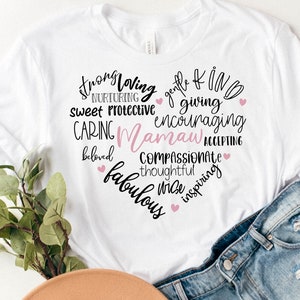 May include: White t-shirt with a heart-shaped design filled with words like "strong," "loving," and "caring." The word "Mamaw" is in pink. Small pink hearts accent the design. The shirt is paired with denim shorts.