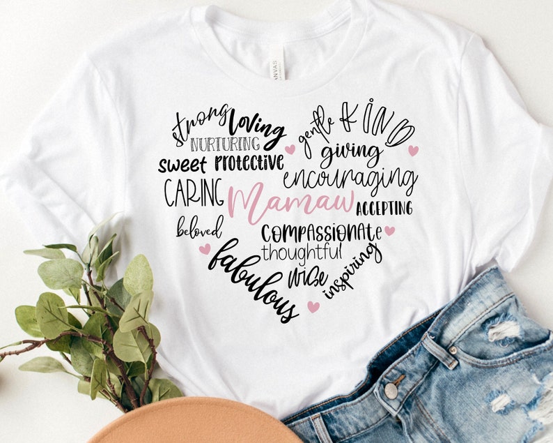 May include: White t-shirt with a heart-shaped design filled with words like "strong," "loving," and "caring." The word "Mamaw" is in pink. Small pink hearts accent the design. The shirt is paired with denim shorts.