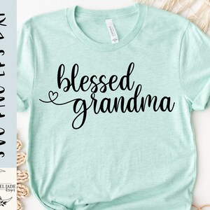 May include: A light blue t-shirt with the text "blessed grandma" in black cursive lettering. A small heart is above the word "grandma".