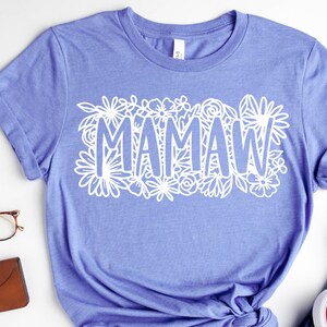 May include: A light purple t-shirt with the word "MAMAW" in white, surrounded by a floral design. The shirt is made of a soft material and has short sleeves. The shirt is a casual wear item.