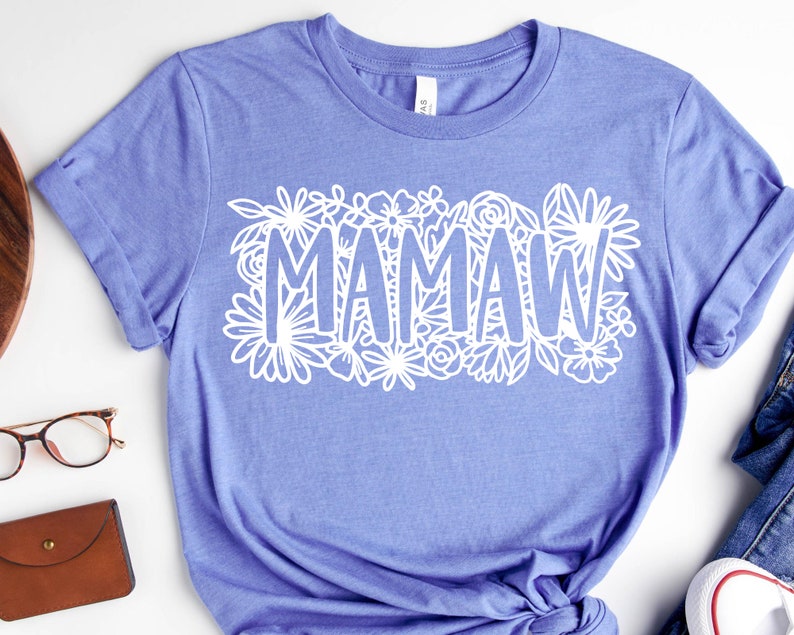 May include: A light purple t-shirt with the word "MAMAW" in white, surrounded by a floral design. The shirt is made of a soft material and has short sleeves. The shirt is a casual wear item.
