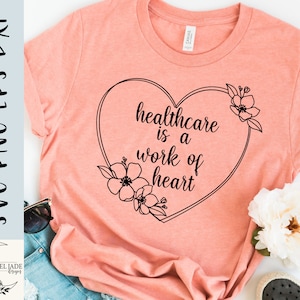 May include: A pink t-shirt with a black heart design that says "healthcare is a work of heart". The heart is surrounded by black floral designs.
