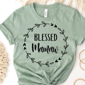 May include: Sage green t-shirt with the words "BLESSED Mamaw" in black script and a black floral wreath design. The shirt is made of a soft material and has rolled-up sleeves.