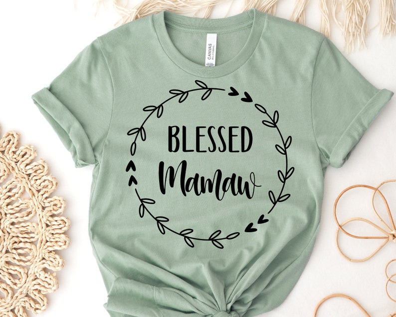 May include: Sage green t-shirt with the words "BLESSED Mamaw" in black script and a black floral wreath design. The shirt is made of a soft material and has rolled-up sleeves.