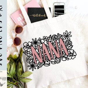 May include: A white tote bag with a black floral design and the word "NANA" in pink lettering. The bag is lying on a white surface with other items, including a notebook and a phone.