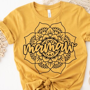 May include: Mustard yellow t-shirt featuring a black mandala design with the word "mamaw" in a cursive font. The shirt is made of soft material and has short sleeves. The design is centered on the front of the shirt.