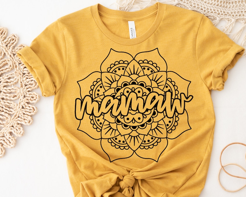 May include: Mustard yellow t-shirt featuring a black mandala design with the word "mamaw" in a cursive font. The shirt is made of soft material and has short sleeves. The design is centered on the front of the shirt.