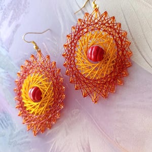 Op de afbeelding: Een paar ovaalvormige hangende oorbellen met ingewikkelde gele en rode draadkunstpatronen. Elke oorbel is in het midden voorzien van een kleine, ronde, rood-wit gestreepte kraal en hangt aan een goudkleurige metalen haak.