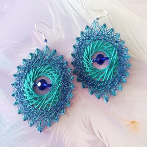 Op de afbeelding: Een paar hangende oorbellen met een geometrisch mandala-ontwerp, gemaakt van blauwgroene en donkerblauwe draad gewikkeld rond een frame. Elke oorbel is in het midden voorzien van een gefacetteerde donkerblauwe kraal en heeft zilverkleurige haken.