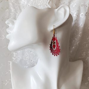 May include: A teardrop-shaped earring featuring a red and dark red thread-wrapped frame with a gold-toned hook. The center of the open frame contains two faceted red beads and one central metallic bronze bead suspended vertically.