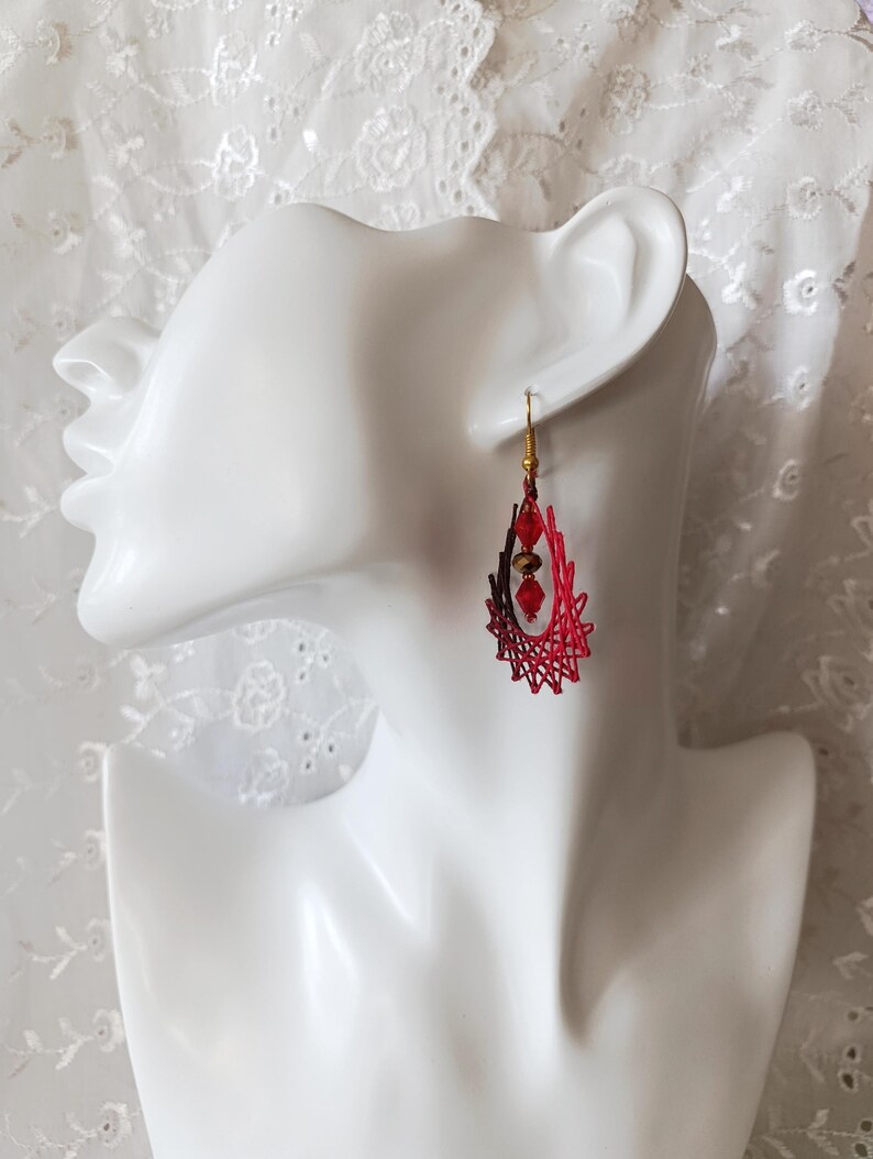 May include: A teardrop-shaped earring featuring a red and dark red thread-wrapped frame with a gold-toned hook. The center of the open frame contains two faceted red beads and one central metallic bronze bead suspended vertically.