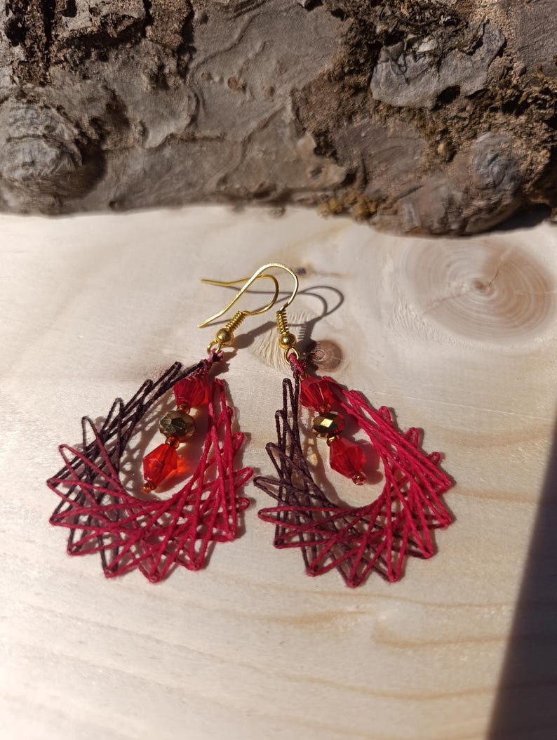 May include: A pair of teardrop-shaped earrings featuring a geometric web of dark red and black thread. Each earring is accented with a vertical arrangement of two red faceted crystal beads and one central metallic gold bead, suspended from gold-tone hooks.