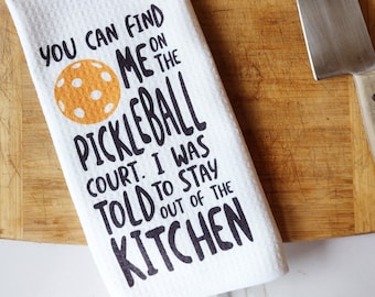 Peut inclure: Une serviette de cuisine blanche avec du texte noir qui dit "You can find me on the pickleball court. I was told to stay out of the kitchen". Une balle de pickleball jaune est représentée à côté du texte.