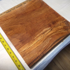 May include: A large piece of wood with a beautiful grain pattern. The wood is a rich brown color with streaks of darker brown and black. The wood is approximately 24 inches wide and 36 inches long. "THREE C'S LUMBER CO." is printed on the wood.