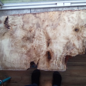 May include: A large, natural wood slab with a unique, swirling grain pattern. The wood is light brown with darker brown streaks and knots. The slab is smooth and polished, and it has a natural, rustic look.