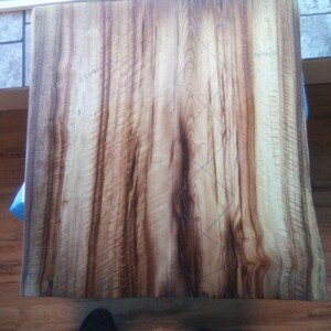 May include: A close-up of a large wooden slab with a natural wood grain pattern. The slab has a light brown color with darker brown streaks. There are pencil markings on the surface of the slab, forming a diamond pattern.