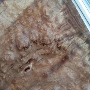 May include: A close-up of a piece of wood veneer with a unique burl pattern. The wood has a light brown color with darker brown streaks and knots. The veneer is smooth and has a natural finish.