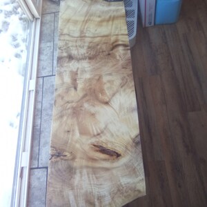 May include: A large, natural wood slab with a unique grain pattern. The wood is light brown with darker brown streaks and knots. The slab is smooth and polished.