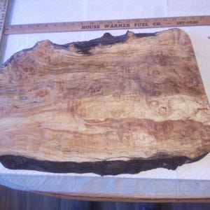 May include: A live edge wooden slab with a natural burl grain pattern in shades of tan, cream, and dark brown. A wooden ruler is positioned along the top edge, showing a length of approximately 18 inches across the top of the wood piece.