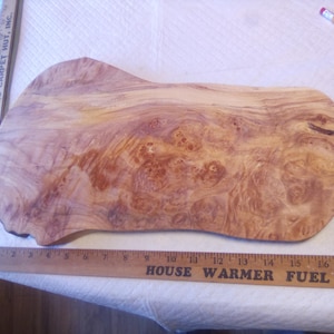 May include: A natural wood cutting board with a live edge and intricate burl grain patterns. A wooden ruler placed along the bottom edge shows the board measures approximately 18 inches in length. The wood features light and dark brown tones.