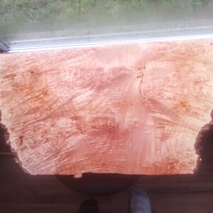 May include: A large piece of wood with a natural, reddish-brown color and a unique grain pattern. The wood has a rough, irregular edge.