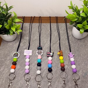 May include: Six lanyards with colourful silicone beads and charms. Each lanyard has a black cord, a metal keyring, and a lobster clasp. Charms include a cat, a duck, and the words "Silly Goose".