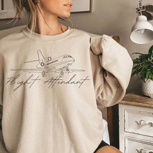Puede incluir: Sudadera beige con cuello redondo y un dibujo lineal negro de un avi&oacute;n y las palabras "Flight Attendant" en una fuente cursiva. La sudadera es de color neutro y tiene mangas largas.