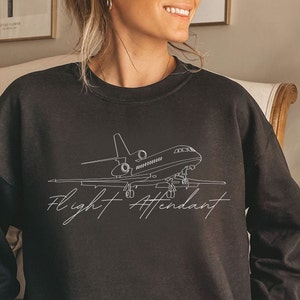 Puede incluir: Sudadera negra con un dibujo lineal blanco de un jet privado y las palabras "Flight Attendant" en cursiva. La sudadera es de cuello redondo.