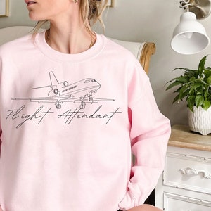 Puede incluir: Sudadera rosa claro con un dibujo lineal negro de un avi&oacute;n y las palabras "Flight Attendant" en fuente cursiva. La sudadera es de cuello redondo.