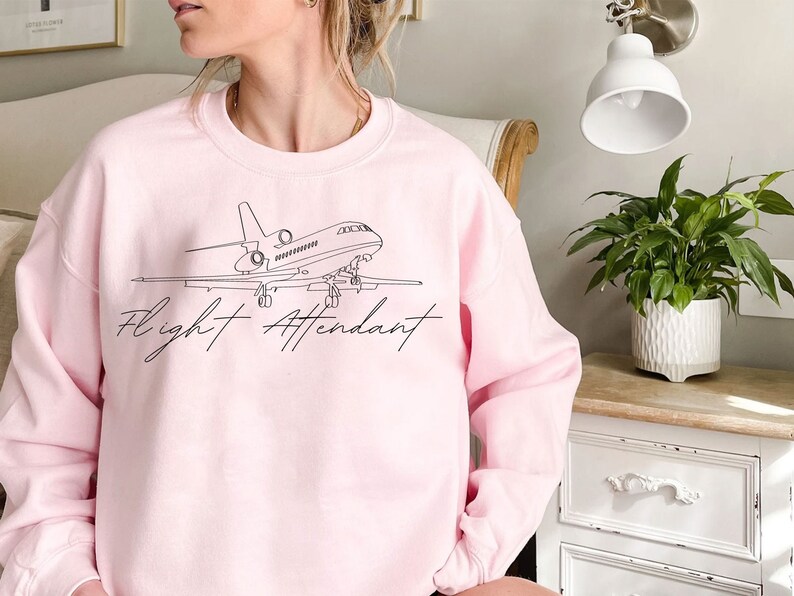 Puede incluir: Sudadera rosa claro con un dibujo lineal negro de un avi&oacute;n y las palabras "Flight Attendant" en fuente cursiva. La sudadera es de cuello redondo.
