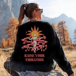 Pode incluir: Camisola de moletom preta com um gráfico retrô de uma mulher em pé na frente de um sol com cogumelos e flores. O texto "RAISE YOUR VIBRATION" é impresso abaixo do gráfico.
