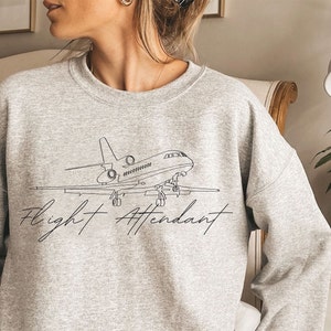 Puede incluir: Una sudadera gris jaspeada con un dibujo lineal negro de un avi&oacute;n y las palabras "Flight Attendant" en cursiva. La sudadera es de cuello redondo.