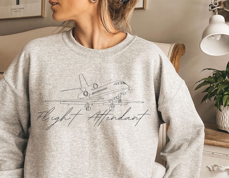 Puede incluir: Una sudadera gris jaspeada con un dibujo lineal negro de un avi&oacute;n y las palabras "Flight Attendant" en cursiva. La sudadera es de cuello redondo.