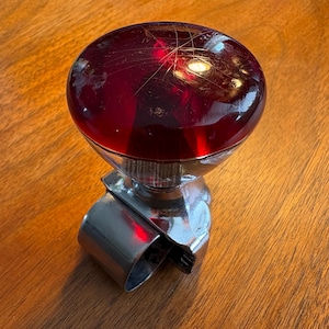 May include: A vintage red glass steering wheel spinner knob with a chrome-colored metal base. The knob is a deep red color and has some scratches on the surface. The metal base has a curved design that wraps around the steering wheel.