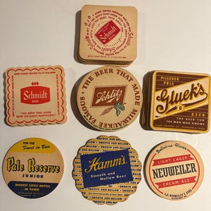 May include: A collection of vintage beer coasters featuring various brands. The coasters are in various shapes, including square and round, and feature logos and text such as "Schmidt Beer", "Schlitz", and "Hamm's".