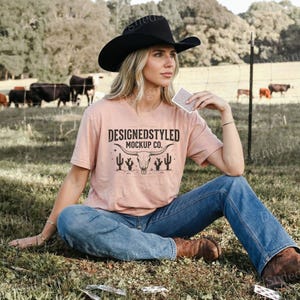 Puede incluir: Una mujer con un sombrero de vaquero negro, una camiseta color melocotón con un diseño gráfico y vaqueros azules. La camiseta presenta el texto "DESIGNEDSTYLED MOCKUP CO." sobre un cráneo de toro y un gráfico de cactus. Sostiene naipes.