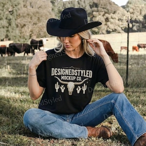 May include: A black t-shirt with the text "DESIGNEDSTYLED MOCKUP CO." and a steer skull graphic. The person is wearing a black cowboy hat and blue jeans. The background includes a grassy field and cattle.