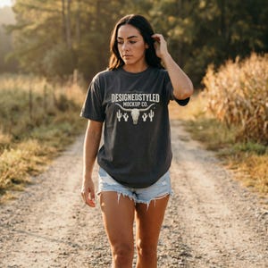May include: A dark gray t-shirt with the text "DESIGNEDSTYLED MOCKUP CO." and a graphic of a bull skull, cacti, and other desert elements. The shirt is worn with denim shorts. The image is taken outdoors.