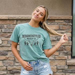 May include: A teal t-shirt with a graphic design featuring a steer skull, cacti, and the text "DESIGNEDSTYLED MOCKUP CO." The shirt is made of a soft material and has short sleeves. The model is wearing denim shorts.