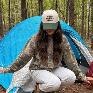 May include: A cream and green baseball cap with the text "YOUR DESIGN HERE". The cap is worn in a forest setting. The person is wearing a camouflage hoodie and grey sweatpants. A blue tent is visible in the background.