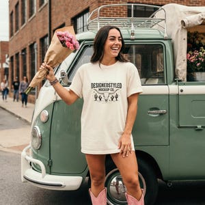 May include: A cream-colored t-shirt with the text "DESIGNEDSTYLED MOCKUP CO." and a graphic of a bull skull and cacti. The person is wearing pink cowboy boots and holding a bouquet of pink flowers. A vintage green and white van is in the background.