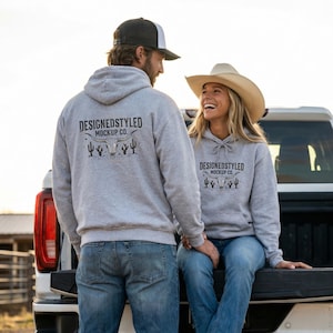 May include: Two people wearing gray hoodies with the text "DESIGNEDSTYLED MOCKUP CO." and a graphic of a steer skull, cacti, and other desert elements. The person on the right is wearing a cowboy hat. They are standing near a truck.