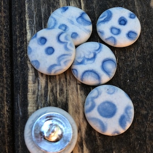Puede incluir: Cinco botones de cerámica blanca con diseños abstractos azules. Los botones son redondos y tienen un acabado liso y brillante.