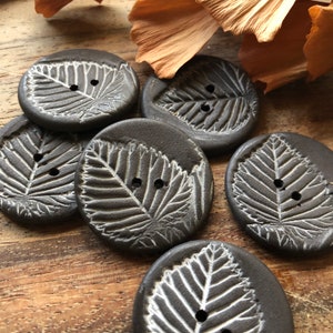 May include: Six dark brown ceramic buttons with a white leaf design. The buttons have two holes each.