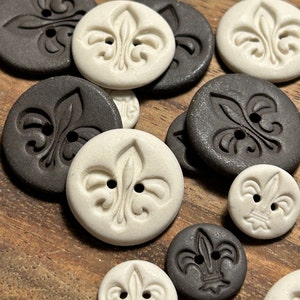 Może przedstawiać: Kolekcja białych i brązowych ceramicznych guzików z motywem lilii. Guziki są ułożone na drewnianej powierzchni.
