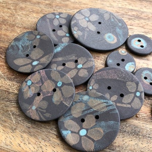 May include: A collection of nine ceramic buttons with a dark brown background and a floral pattern in shades of brown, gold, and blue. The buttons have two holes each.