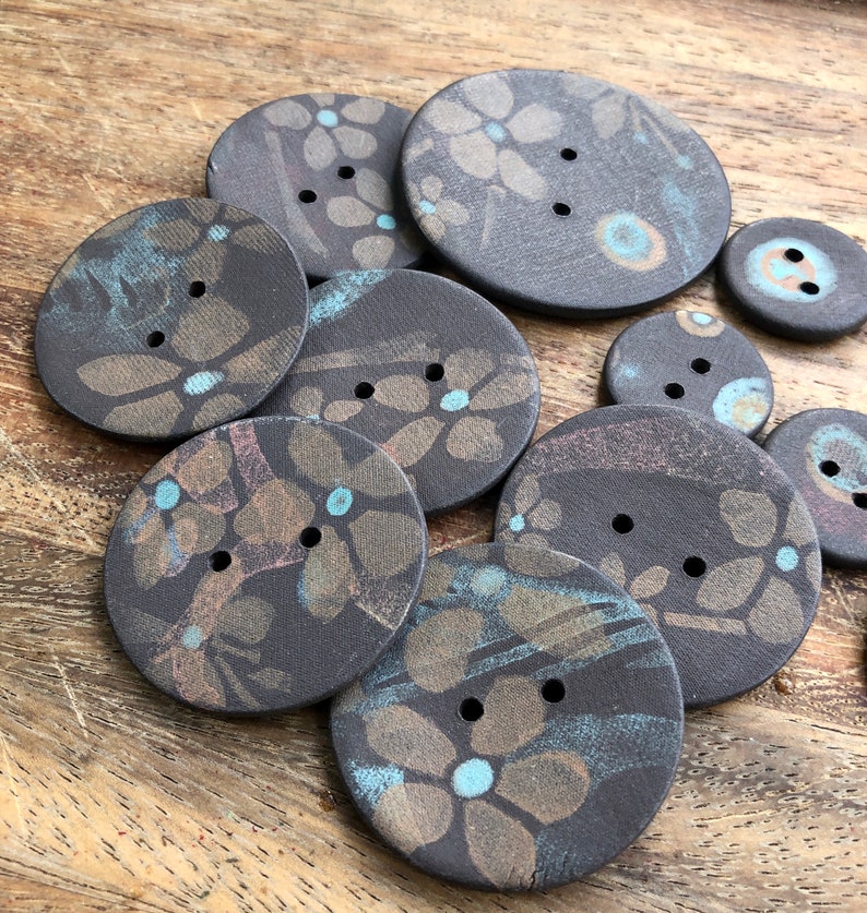 May include: A collection of nine ceramic buttons with a dark brown background and a floral pattern in shades of brown, gold, and blue. The buttons have two holes each.